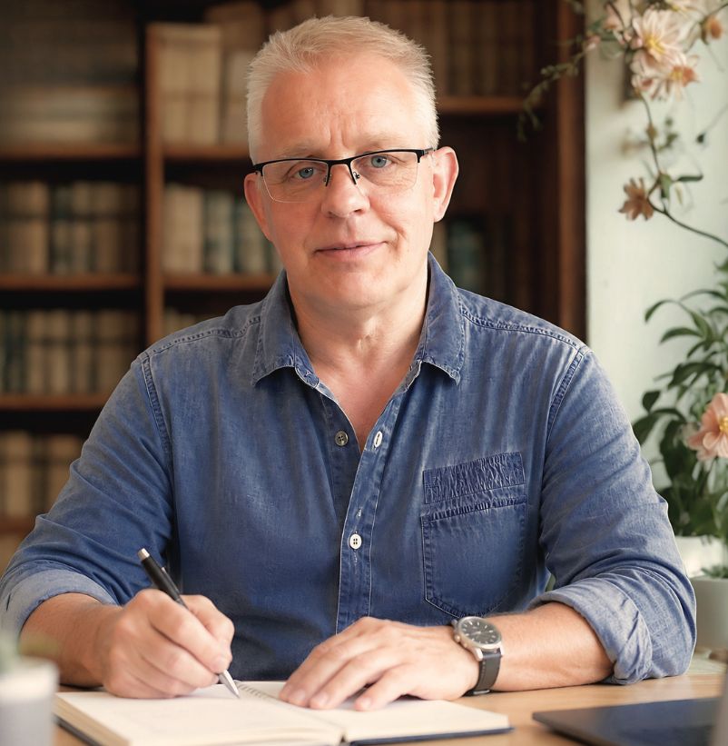 Man with glasses in a denim shirt signs a notebook at a desk, bookshelf behind him.